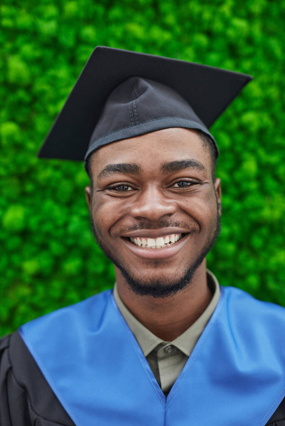 Κάθετη γκρο πλαν πορτρέτο του ευτυχισμένου Αφροαμερικανού άνδρα φορώντας καπέλο αποφοίτησης και χαμογελώντας στην κάμερα, ενώ στέκεται πάνω σε πράσινο φόντο φυτό - Φωτογραφία, εικόνα