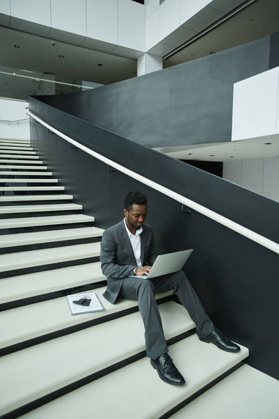 Retrato gráfico de larga duración de un hombre de negocios afroamericano usando una computadora portátil mientras está sentado en las escaleras en el pasillo de la oficina - Foto, imagen