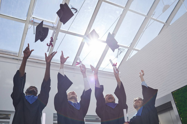 Visão de baixo ângulo em grupo diversificado de graduados universitários jogando chapéus no ar contra o céu com luz solar, espaço de cópia - Foto, Imagem