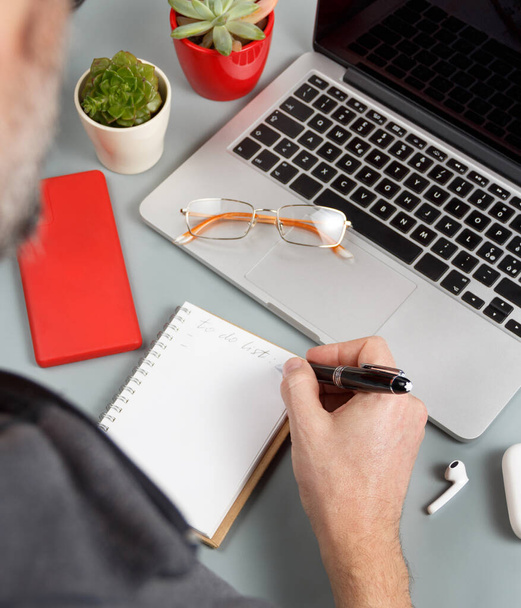 Homem escrevendo PARA FAZER LISTA perto de laptop em uma mesa de escritório cinza de perto - Foto, Imagem