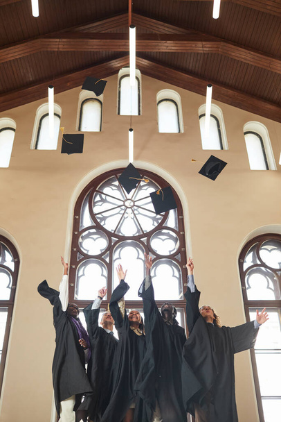Вертикальный широкоугольный вид на трех молодых людей, бросающих люки в воздух во время выпускной церемонии в помещении классического школьного аудитории, копировальное пространство - Фото, изображение