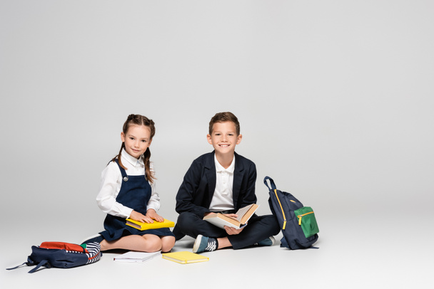 happy schoolkids in prestigious uniform sitting with backpacks and books on grey - Фото, изображение