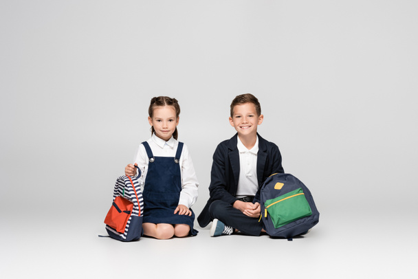 cheerful schoolkids in uniform sitting with backpacks on grey - Zdjęcie, obraz