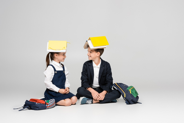 happy schoolkids in uniform with books on heads sitting and looking at each other on grey - Zdjęcie, obraz