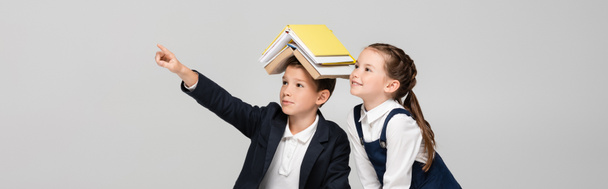 uczeń z książkami na głowie wskazując palcem w pobliżu wesołej dziewczyny odizolowany na szarości, sztandar - Zdjęcie, obraz