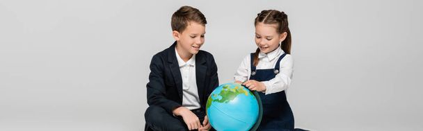 glückliches Schulmädchen und Schuljunge mit Blick auf Globus isoliert auf grauem Banner - Foto, Bild