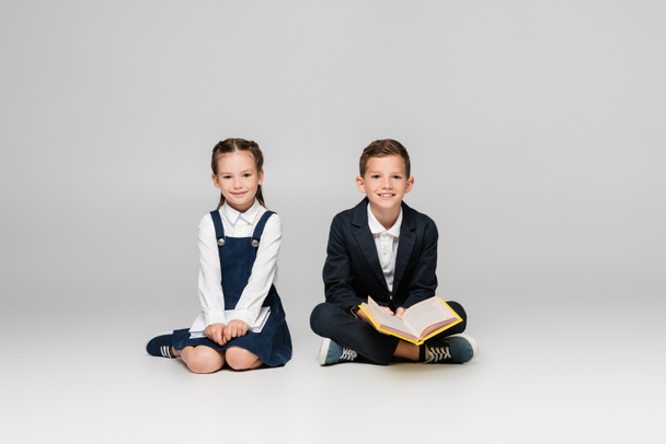 pozytywne dzieci w wieku szkolnym siedzące z książkami i uśmiechnięte na szaro - Zdjęcie, obraz
