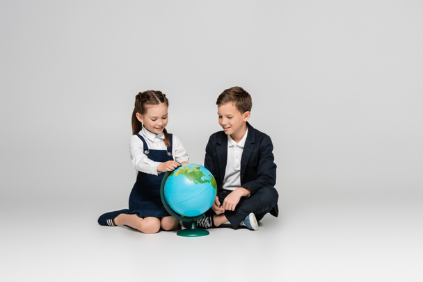 happy schoolgirl and schoolboy looking at globe while sitting on grey - Zdjęcie, obraz