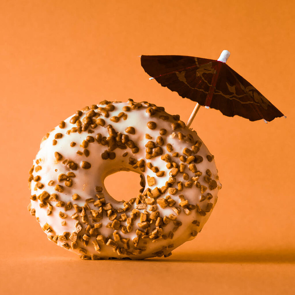 アーモンドトッピングのホワイトチョコレートドーナツとオレンジの背景の装飾傘. - 写真・画像