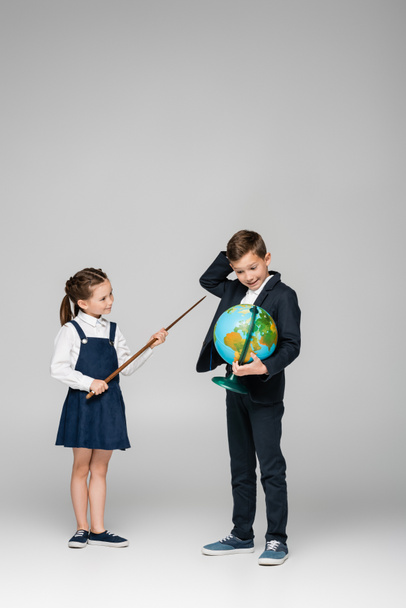 Lächelndes Schulmädchen hält Zeigestöckchen neben verwirrtem Jungen mit Globus auf grau  - Foto, Bild