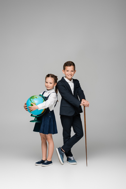 smiling schoolboy holding pointing stick near girl in dress with globe on grey  - Fotoğraf, Görsel