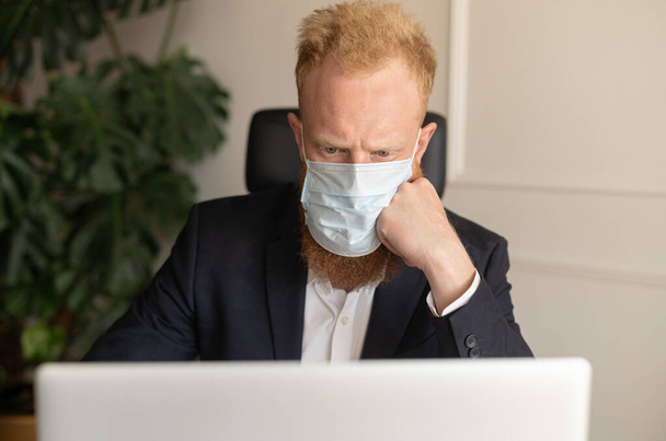 Thoughtful red-haired businessman in smart casual suit wearing protective face mask - Fotoğraf, Görsel