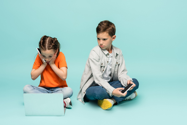 shocked girl talking on smartphone and using laptop near sad boy on blue - Φωτογραφία, εικόνα
