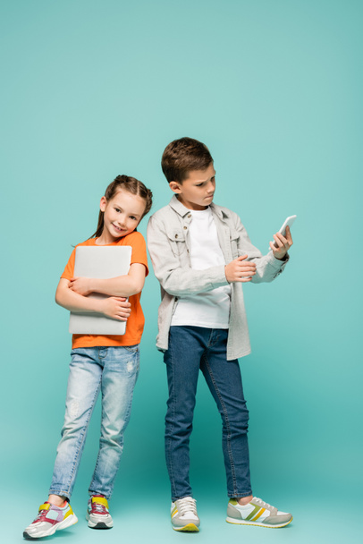 happy girl with laptop standing near boy using smartphone on blue - Photo, Image