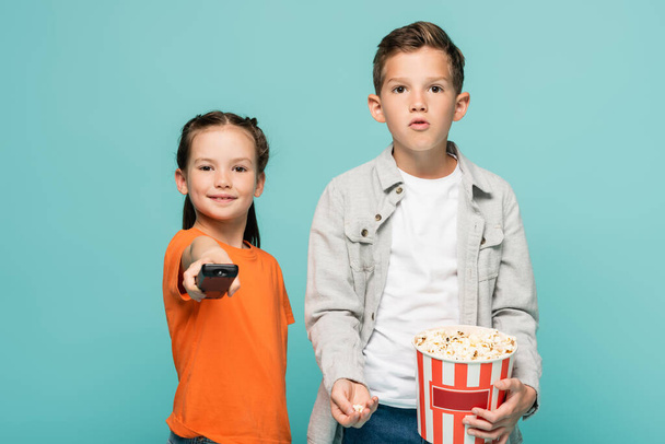 girl holding remote controller near boy with popcorn bucket isolated on blue - Foto, afbeelding