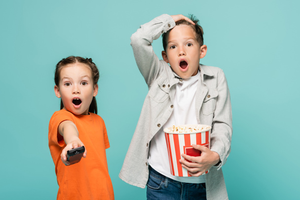 shocked girl holding remote controller near boy with popcorn bucket isolated on blue - Foto, afbeelding