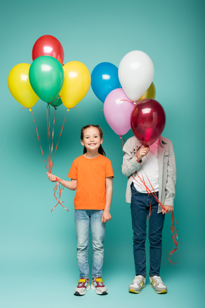 happy girl holding colorful balloons near boy obscuring face on blue  - Zdjęcie, obraz