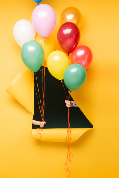 cropped view of kids holding colorful balloons through hole on yellow background - Photo, Image
