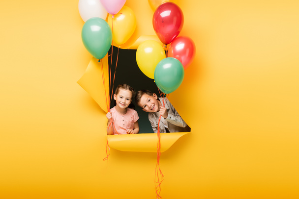 cheerful kids holding colorful balloons through hole on yellow background - Photo, Image