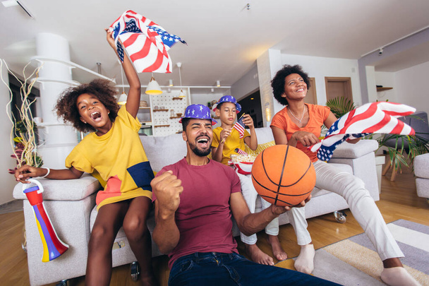 Χαρούμενη Αφρο-αμερικανική οικογένεια βλέποντας τηλεόραση και επευφημίες μπάσκετ παιχνίδια στον καναπέ στο σπίτι. - Φωτογραφία, εικόνα