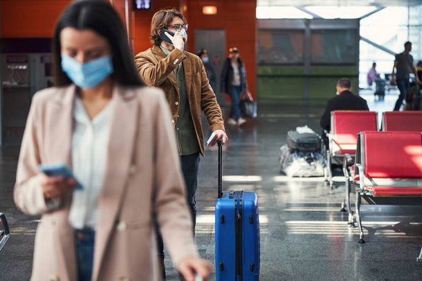 Стильний чоловік в медичній масці розмовляє на мобільному телефоні в аеропорту
 - Фото, зображення