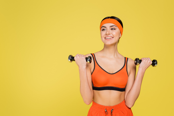 joyful sportswoman looking away while working out with dumbbells isolated on yellow - Foto, Bild