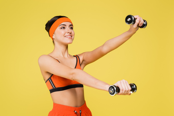smiling sportswoman in orange sportswear training with dumbbells isolated on yellow - Foto, imagen