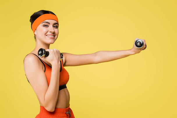 pleased woman smiling at camera while working out with dumbbells isolated on yellow - Fotografie, Obrázek
