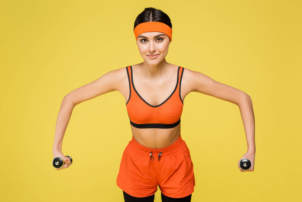 young sportswoman in orange sportswear training with dumbbells isolated on yellow - Фото, зображення