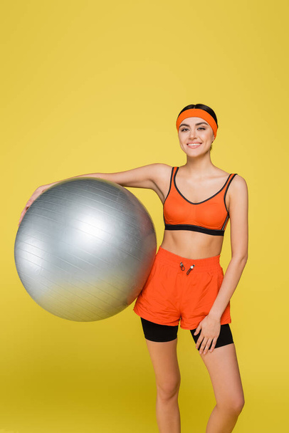 happy sportive woman with silver-grey fitness ball smiling at camera isolated on yellow - Fotoğraf, Görsel