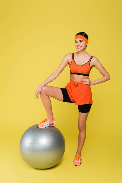 sportive woman stepping on fitness ball while standing with hand on hip on yellow background - Fotografie, Obrázek