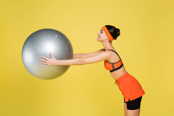 side view of smiling sportswoman exercising with fitness ball isolated on yellow - Фото, изображение