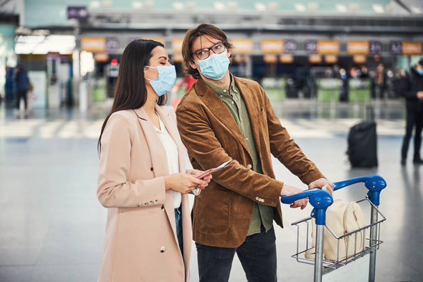 Schönes Paar in medizinischen Masken läuft Flughafen-Terminal hinunter - Foto, Bild