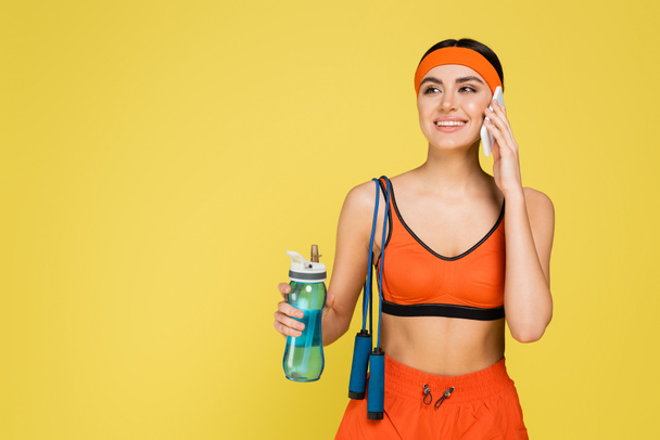 smiling woman with sports bottle and jump rope calling on smartphone isolated on yellow - Фото, изображение