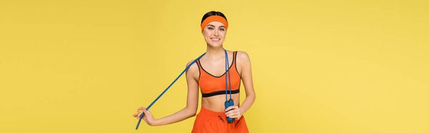 örömteli sportoló nő ugrókötéllel mosolyog a kamera elszigetelt sárga, banner - Fotó, kép