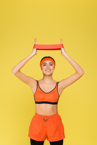 cheerful woman in orange sportswear training with resistance band isolated on yellow - Фото, изображение