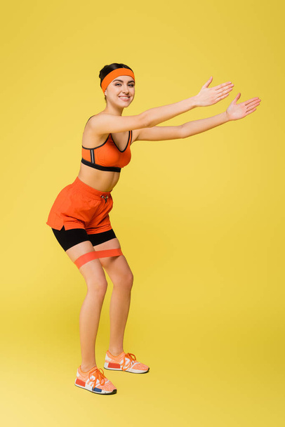 joyful sportswoman looking at camera while exercising with elastics isolated on yellow - Photo, Image