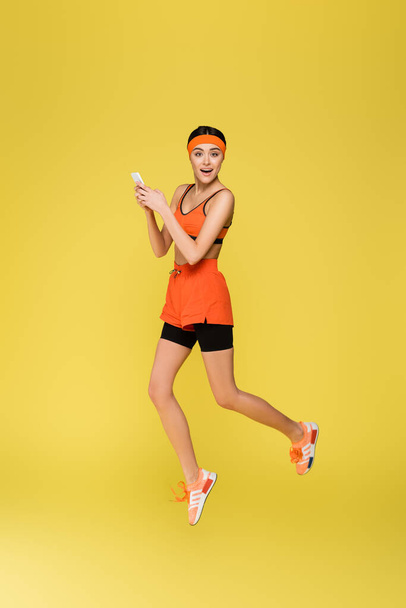 young sportswoman jumping while using smartphone isolated on yellow - Fotoğraf, Görsel