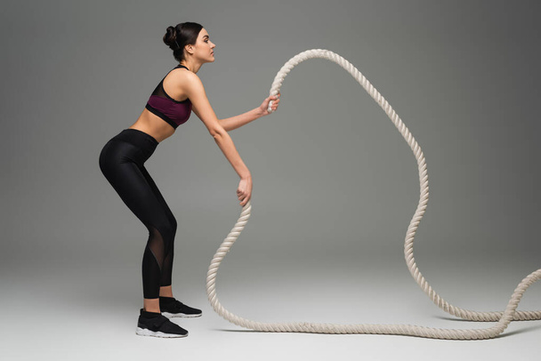 side view of sportswoman training with battle ropes on grey background - Fotoğraf, Görsel