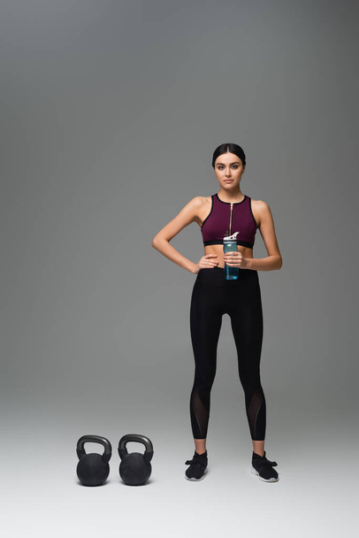 confident woman with sports bottle standing near kettlebells on grey background - Foto, imagen