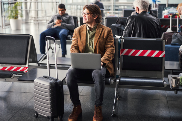 Joyful young man using laptop in departure lounge - Photo, Image