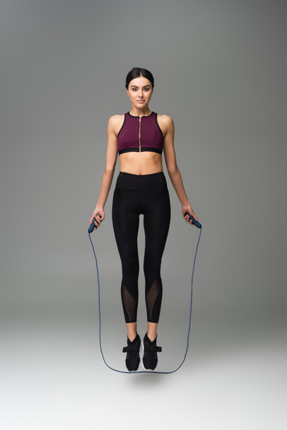 sportswoman looking at camera while jumping with skipping rope on grey background - Photo, Image