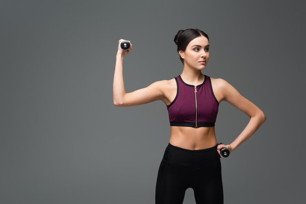 pretty sportive woman looking away while exercising with dumbbells isolated on grey - Foto, imagen