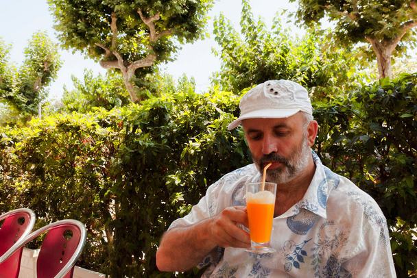 Egy érett férfi szürke szakállal baseball sapkában ül egy sötét kávézóban és narancslevet iszik. - Fotó, kép