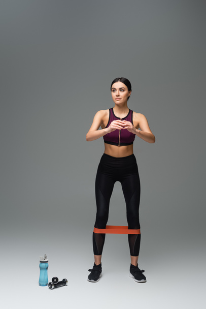 full length view of sportive woman looking away while training with elastics on grey background - Photo, Image