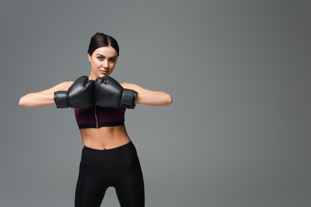 young sportswoman in boxing gloves looking at camera isolated on grey - Фото, изображение