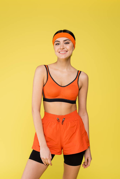 young woman in orange sportswear smiling at camera isolated on yellow - Foto, imagen