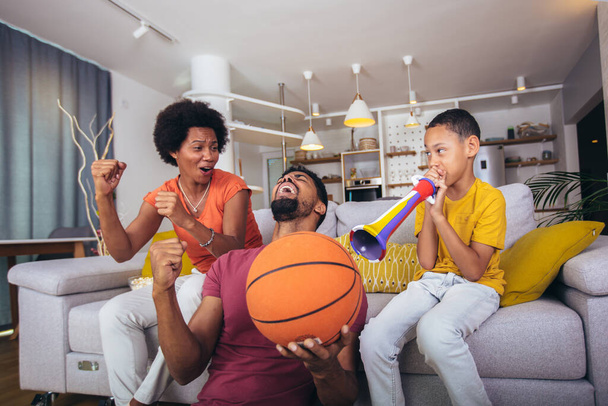 Feliz familia afroamericana viendo la televisión y animando los juegos de baloncesto en el sofá en casa. - Foto, imagen