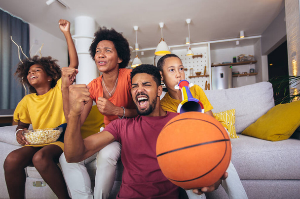 Boldog afro-amerikai család néz TV-t és szurkolás kosárlabda játékok kanapén otthon. - Fotó, kép