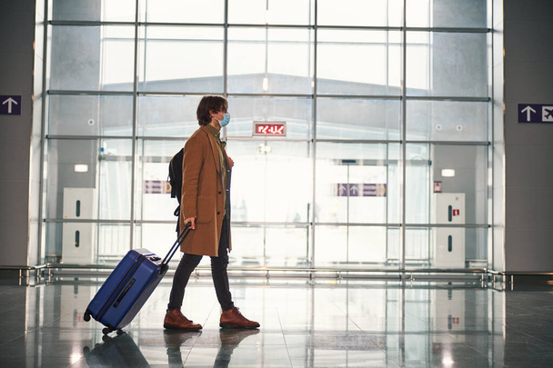 Κομψός νεαρός άνδρας με ιατρική μάσκα που μεταφέρει ταξιδιωτική βαλίτσα στο αεροδρόμιο - Φωτογραφία, εικόνα